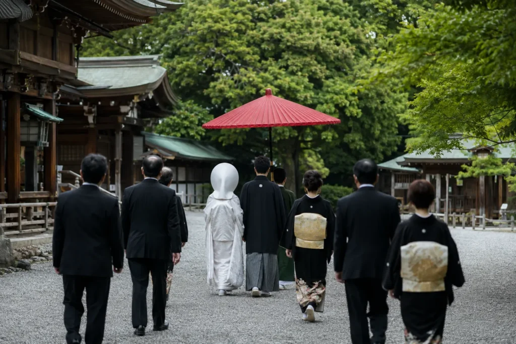 神社の砂利道を、白無垢に綿帽子の新婦と紋付羽織袴の新郎が赤い和傘の下を歩き、黒留袖と礼服の親族が続いている