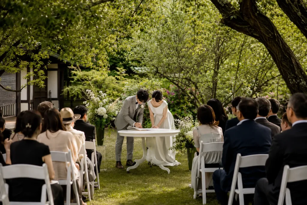 木々に囲まれた芝生の会場で、白いテーブルに置いた結婚証明書に署名する新郎新婦。周囲を白い椅子に座ったゲストが囲んでいる