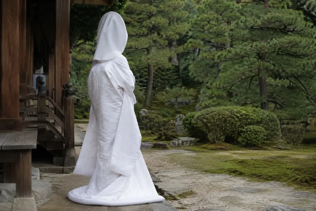 日本庭園に面した木造の建物の前に立つ、白無垢に綿帽子姿の花嫁の後ろ姿。打掛から掛下、小物まですべて白でそろえられている